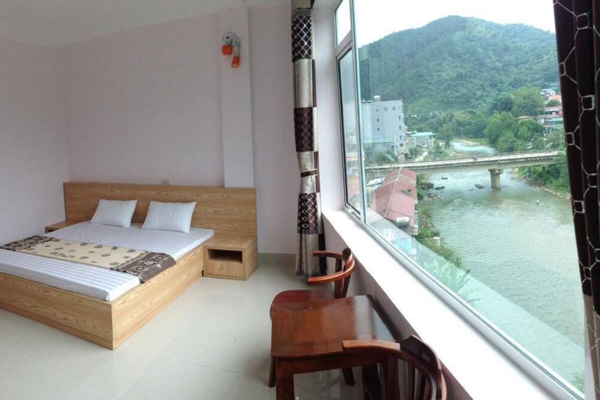 Un dormitorio con una cama de madera, un taburete pequeño de madera, una mesa y vistas a un río y un puente.