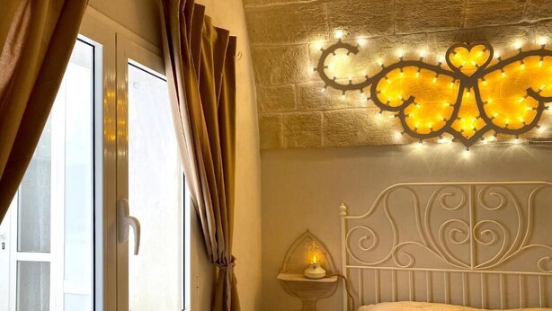 A bedroom with a bed, a window with curtains, a wall-mounted light fixture, and a stone wall. - The Pink Pumo (Monopoli)