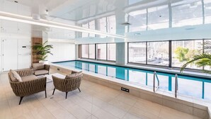 A modern indoor pool area with clear glass walls, a lounge with cushioned seating, and a visible depth marker.