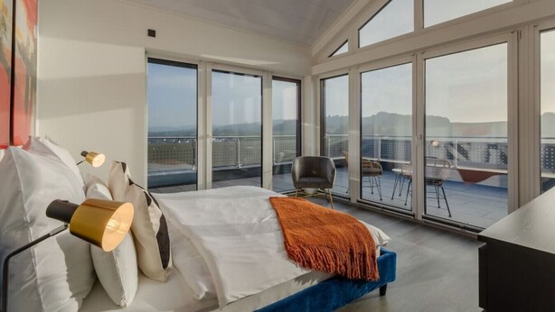 A modern bedroom with a large bed, a view of the city through floor-to-ceiling windows, and a balcony with outdoor seating. - Modern Oasis w/ HotTub, View, Gym (Oberbettingen)