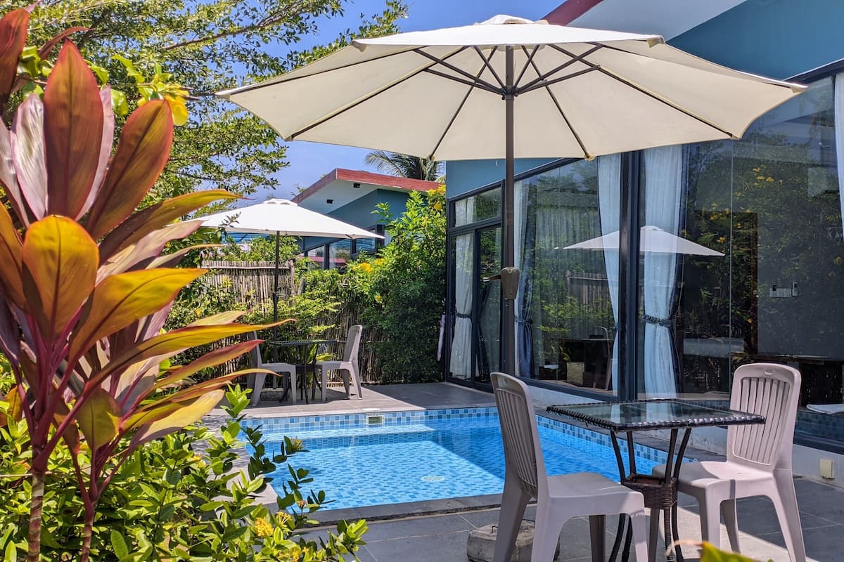 A poolside area with a glass table and chairs, an umbrella, and a view of the surrounding greenery and buildings.