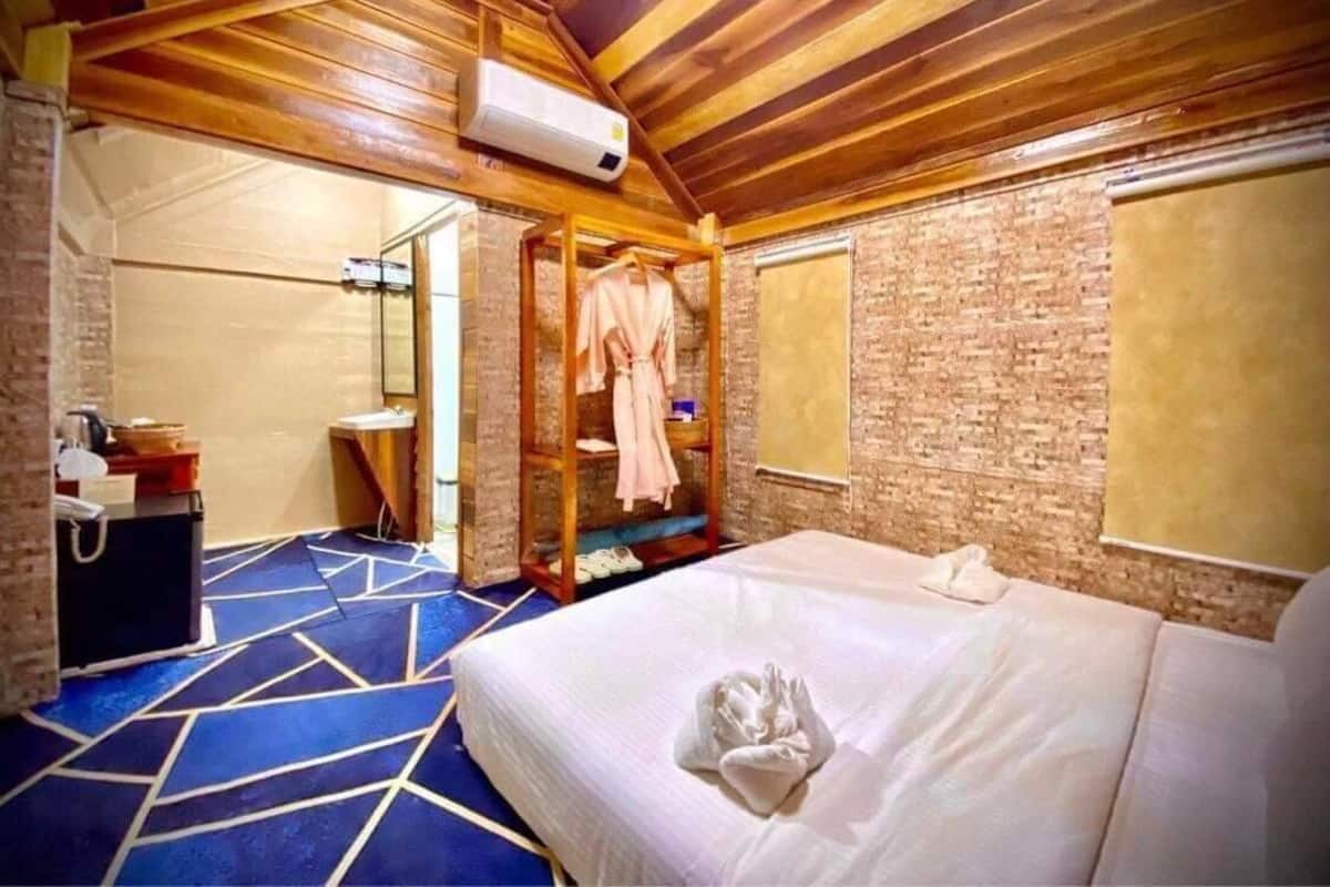 A hotel room with a wooden ceiling, a brick wall, a bed with white linens, a robe rack, and a small desk with a telephone.