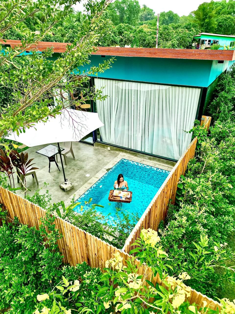 A swimming pool with a person sitting on a float, surrounded by lush greenery and a wooden fence.