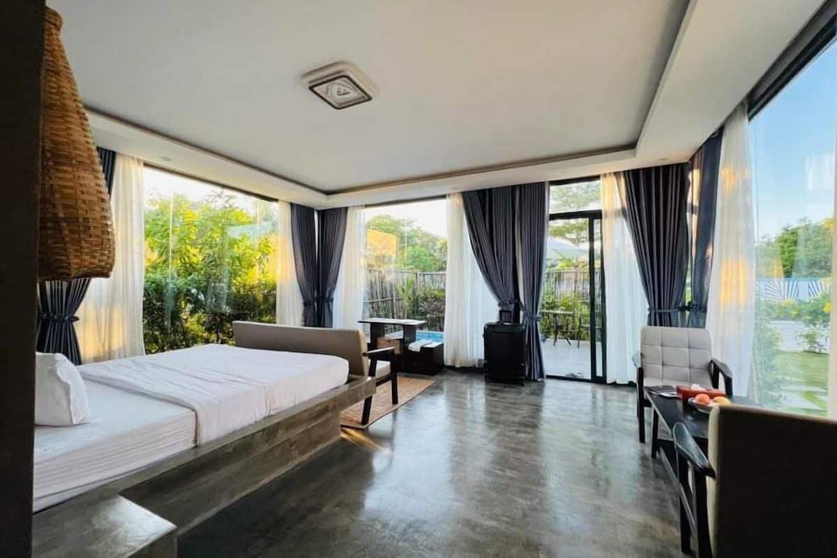 A modern hotel room with a bed, a small table, a chair, and a view of greenery outside.