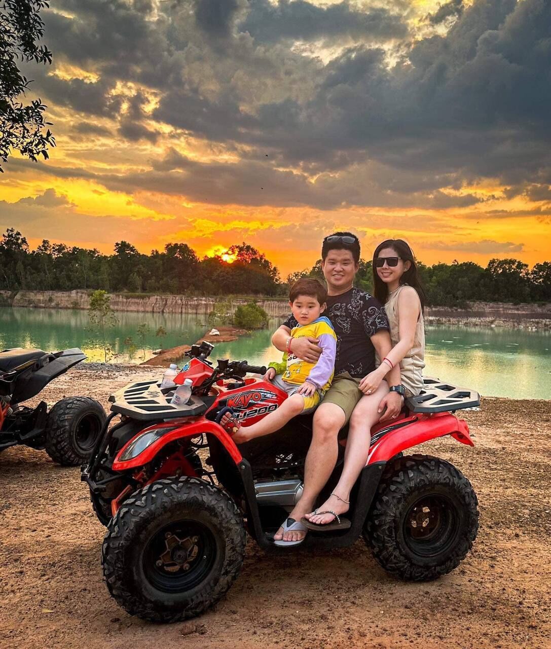 A family on a red ATV near a body of water with a sunset in the background.