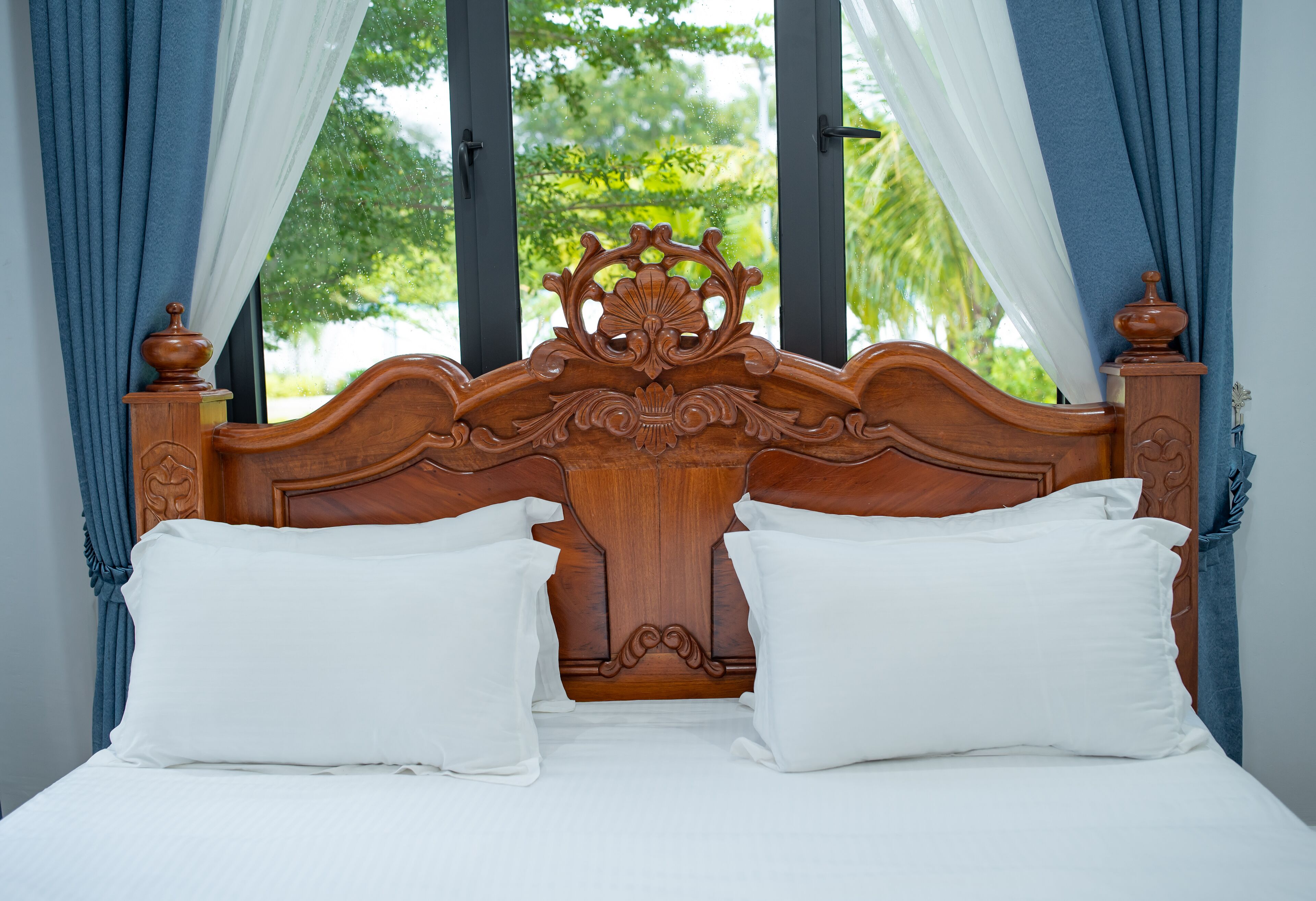 A wooden bed with white pillows and curtains, a window with a view of greenery.