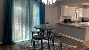 Un comedor con una mesa redonda y cuatro sillas, una araña y una cocina con microondas y fregadero.