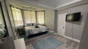 A bedroom with a bed, a TV mounted on the wall, a door, and a window with curtains. - Roomy & Newly Renovated Guest House, Kitchen, Desk, Pet Friendly, Shared Patio! (Ellenwood)