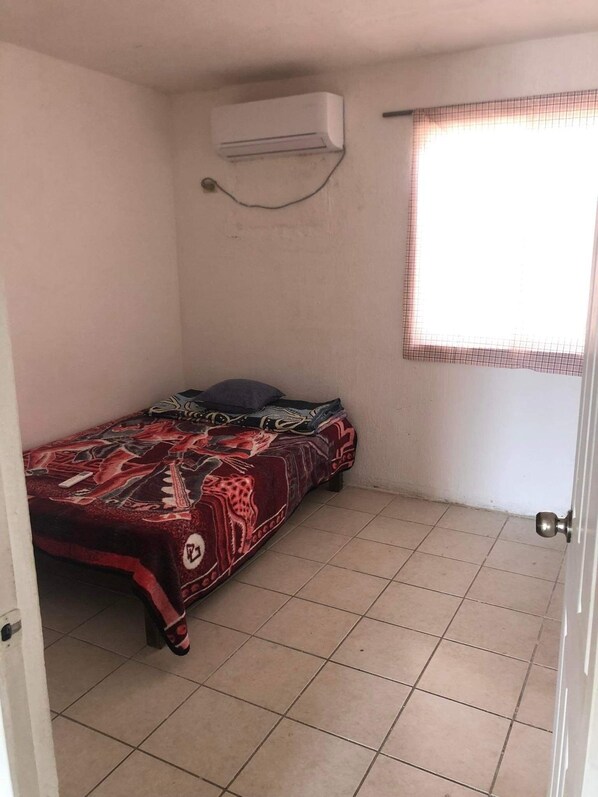 A small, single-bed room with a patterned bedspread, an air conditioning unit mounted on the wall, and a window with a checkered blind.
