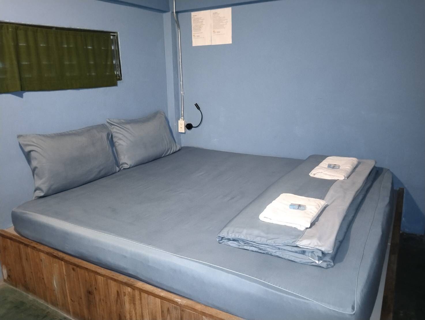 A single bed with a grey mattress and two pillows, a wooden headboard, a power outlet, and a window with blinds.