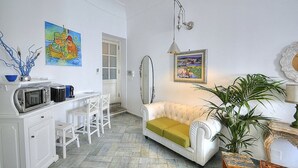 A compact living space with a kitchenette, a white sofa with a green cushion, a dining area with a white chair, and a large potted plant. - Suite Baldea: An elegant and welcoming apartment that is part of an ancient watch tower built sheer above the sea, with Free WI-FI. (Positano)