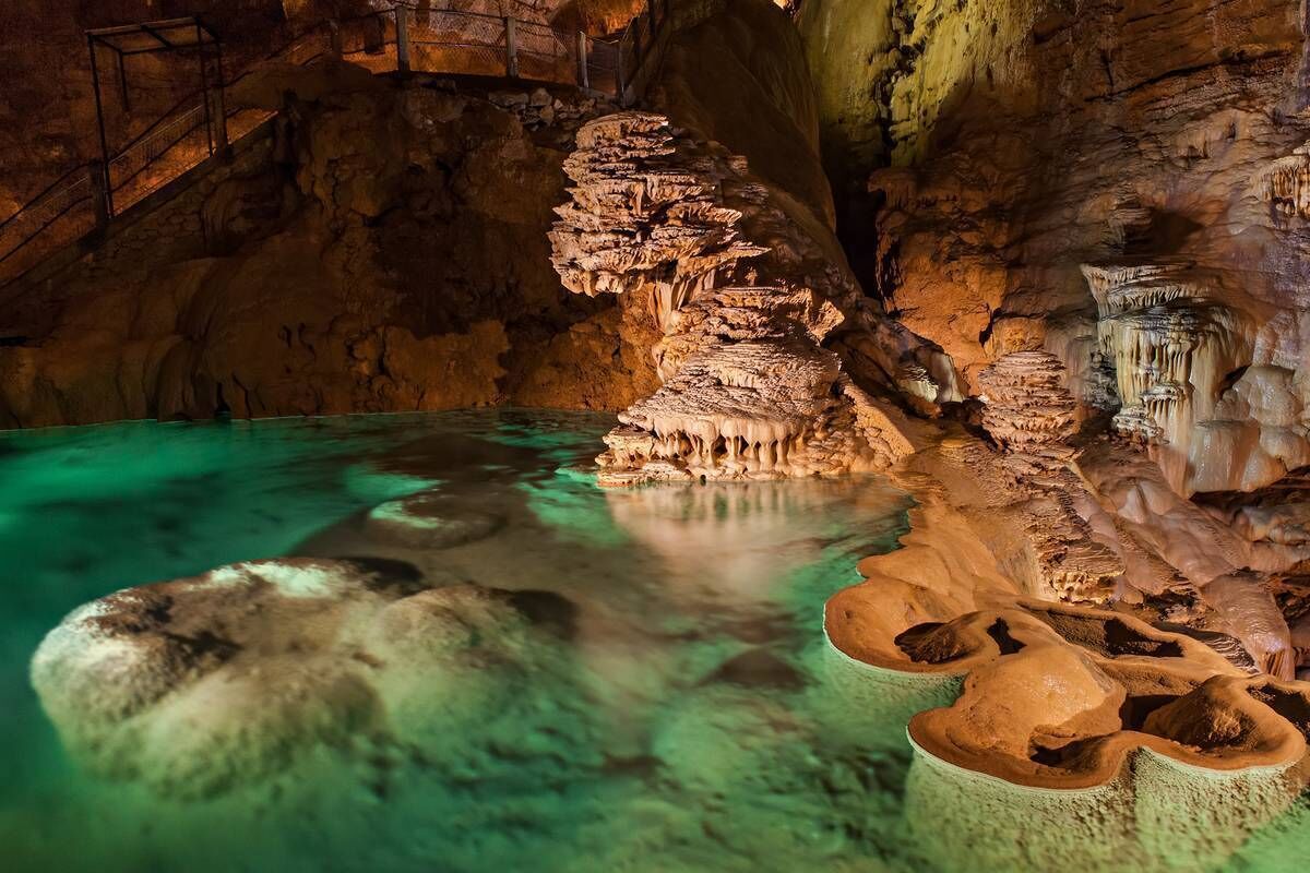 Une grotte ornée de stalactites et de stalagmites, une mare d’eau et un escalier.
