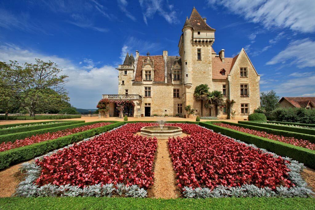 A large, historic building with a symmetrical garden featuring a central fountain, surrounded by manicured hedges and flower beds.