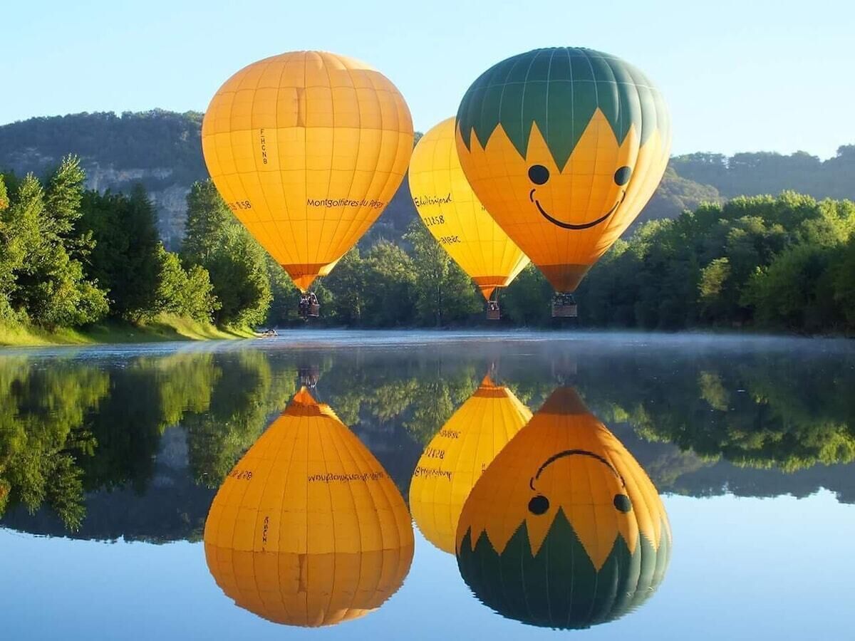 A group of hot air balloons over a calm lake with reflections, surrounded by trees and hills.