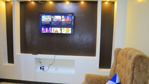 Ein Wohnzimmer mit flachbildfernseher an der Wand, einem braunen Sessel mit blauem und gelbem Kissen und einem kleinen Teppich auf dem Boden.