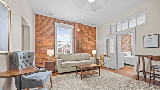 A living room with a brick wall, a sofa, a coffee table, and a patterned rug. - 30-Night Minimum South Main Apartment (Saint Charles)