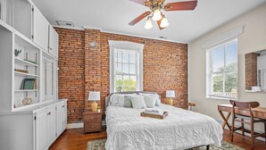 A bedroom with a brick wall, a bed with white bedding, a desk, and a chair. - 30-Night Minimum South Main Apartment (Saint Charles)