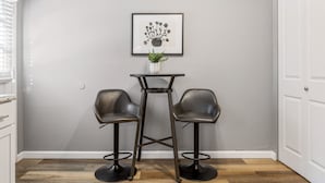Ein Eckzimmer mit grauer Wand, einem gerahmten Blumenbild, einem kleinen Tisch mit Topfpflanze und zwei ledernen Barhocker.