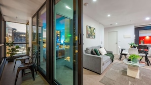 A modern living room with a sofa, dining table, and chairs, a glass door leading to a city view, and a kitchen area with red lighting. - Lovely City Escape 2B2B Unit in Melbourne Central (Melbourne)