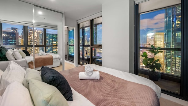 A modern hotel room with a large bed, a sofa, and a city view through floor-to-ceiling windows. - Lovely City Escape 2B2B Unit in Melbourne Central (Melbourne)