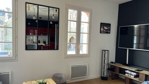 Un salon moderne avec un téléviseur, un plancher en bois, une table à manger agrémentée d’une plante, une grande fenêtre donnant sur des immeubles et un mur d’accent de couleur noire.