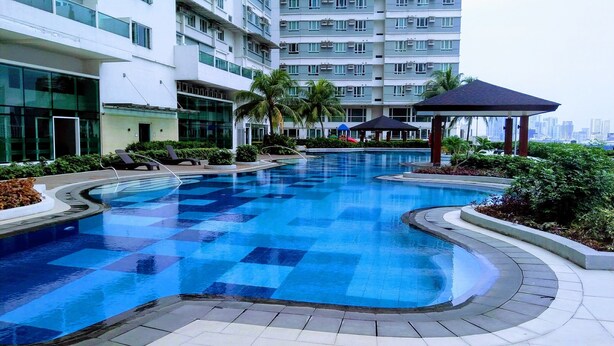 A swimming pool with a checkered pattern, surrounded by lounge chairs and palm trees, with a modern building in the background. - Dimacs Staycation  (Makati)