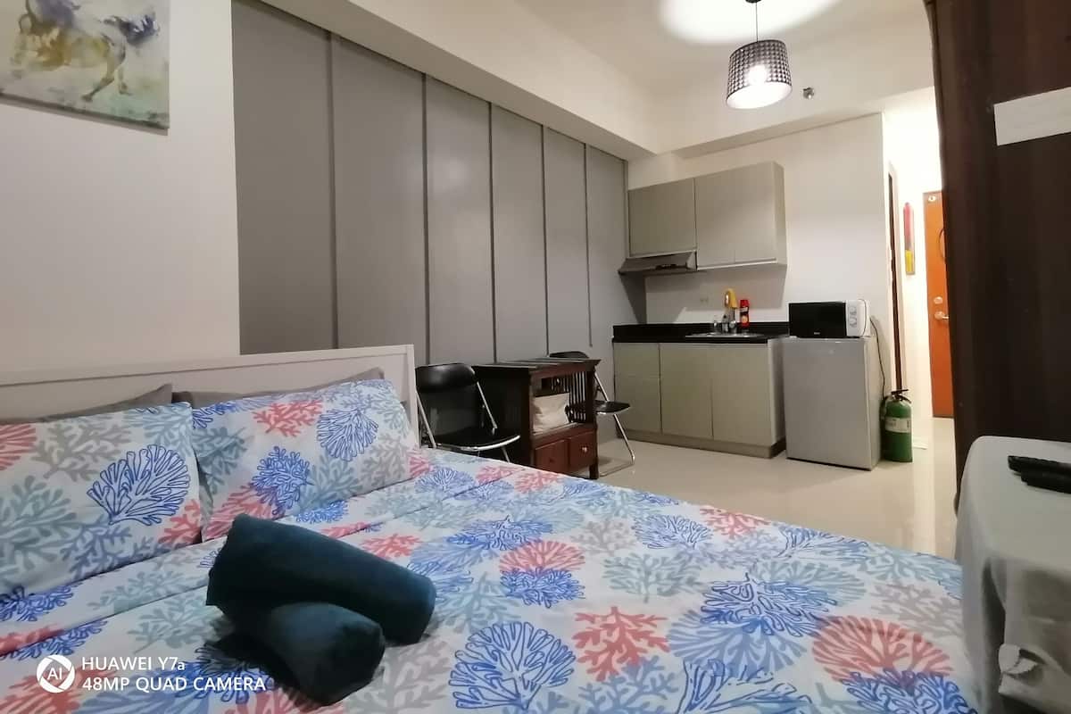A bedroom with a bed, a chair, a desk, a kitchenette, and a bathroom.