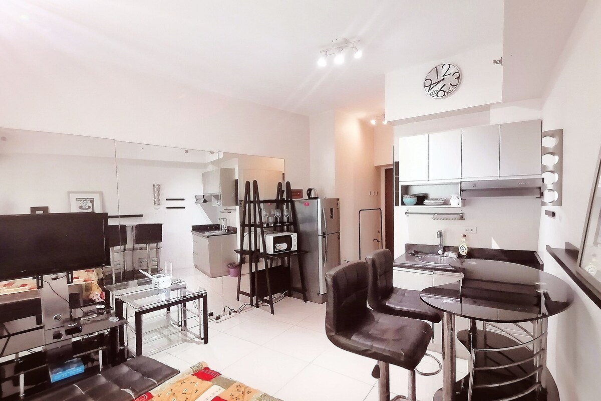 A modern kitchen with white cabinets, a stainless steel refrigerator, and a microwave. There is a dining area with a black chair and a small table.