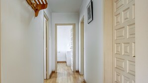 Um corredor estreito com piso de madeira e paredes brancas. Há uma porta com painel decorativo à direita e uma porta com design simples à esquerda.