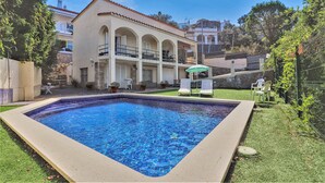 Piscina com deck ladrilhado, rodeada por um gramado bem cuidado e um edifício com telhado em azulejos e várias varandas.