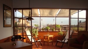 Een zonnig terras met een tafel en stoelen, potplanten en een uitzicht op een stadsbeeld door grote ramen.