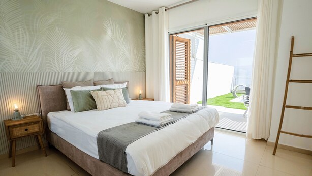Une chambre spacieuse avec un grand lit, une table de chevet, une échelle en bois et une vue sur une pelouse verdoyante, visible à travers une porte-fenêtre coulissante.