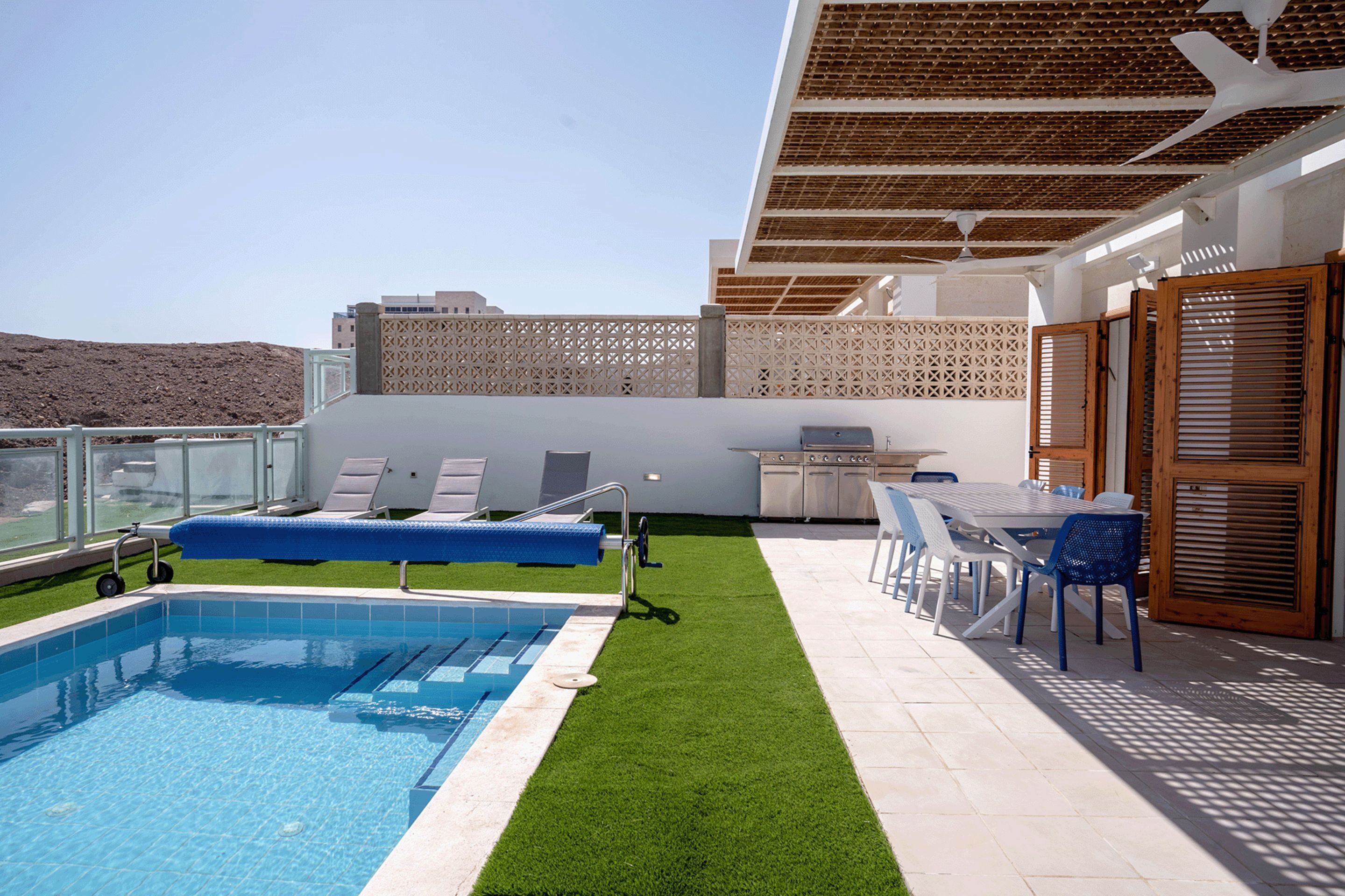 A rooftop pool area with a swimming pool, sun loungers, a table with chairs, and a grill.