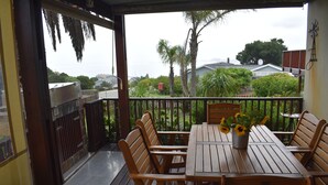 A wooden deck with a table and chairs, a potted plant with sunflowers, and a view of houses and trees in the background. - Brenton Bliss, beach view, walk to beach, safe secluded area on the Garden Route (Knysna)