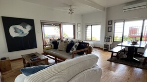 A spacious living room with a large sofa, a coffee table, and a dining area with a table and chairs. There is a ceiling fan, large windows, and a painting on the wall. - Brenton Bliss, beach view, walk to beach, safe secluded area on the Garden Route (Knysna)