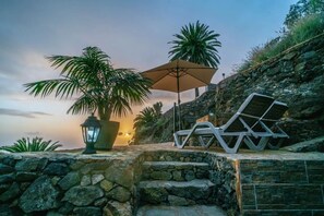 Un sendero de piedra, sillones de descanso, una sombrilla grande y una planta en maceta.