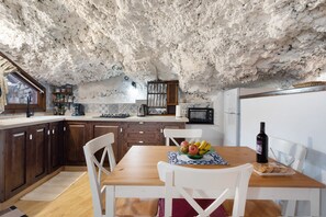A rustic kitchen with wooden cabinets, a dining table with chairs, a fruit bowl, a bottle of wine, and a microwave.