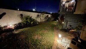 Vue nocturne d'une cour arrière avec un chemin dallé, un mur blanc et quelques plantes.