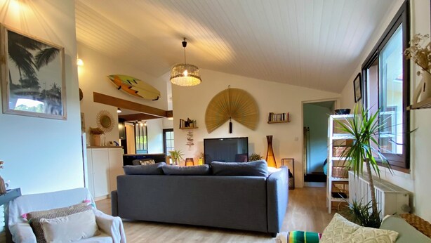Una sala de estar con sofá, sillón y mesa de centro. Hay un televisor, un ventilador y una planta en una estantería.