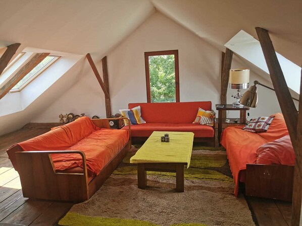 A cozy loft bedroom with a sloped ceiling, wooden beams, a red sofa, a yellow table, and a window with a view of greenery.