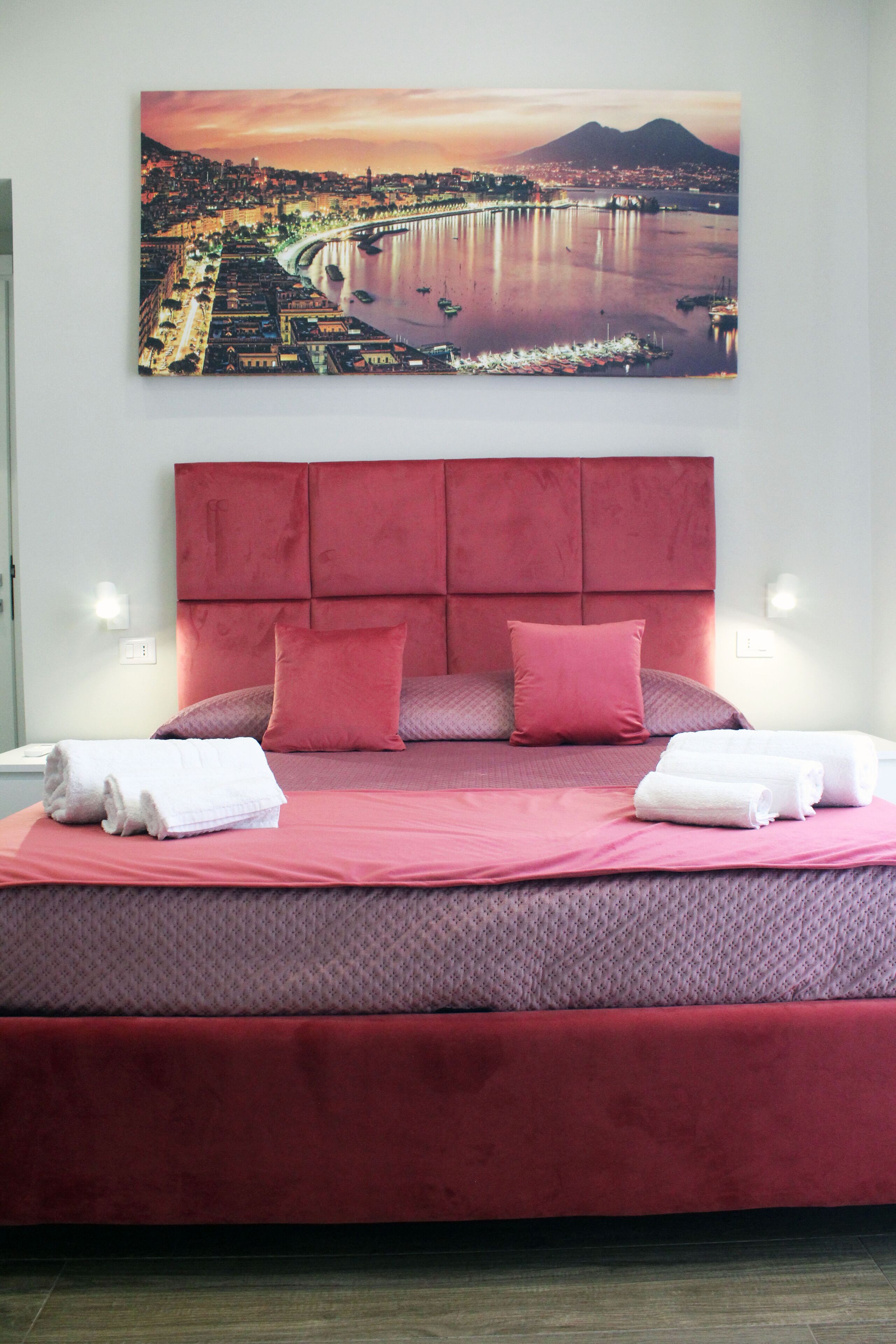 Ein Bett mit rotem Kopfteil und Kissen, darüber ein Stadtbild als Gemälde, und zwei Nachttischlampen.