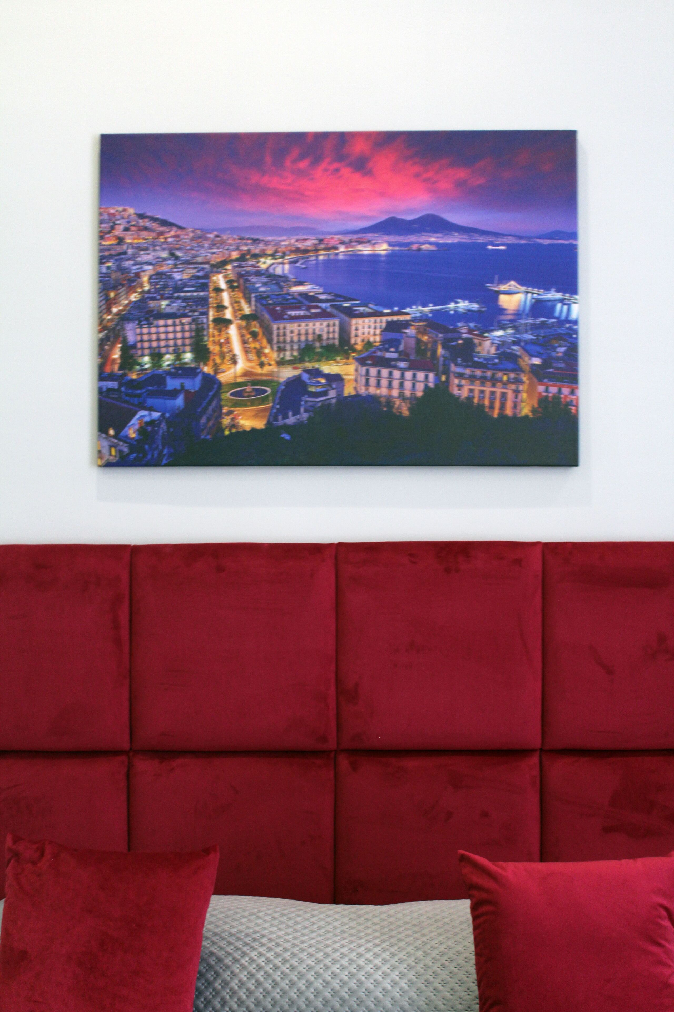 A cityscape painting on a wall above a red upholstered headboard.