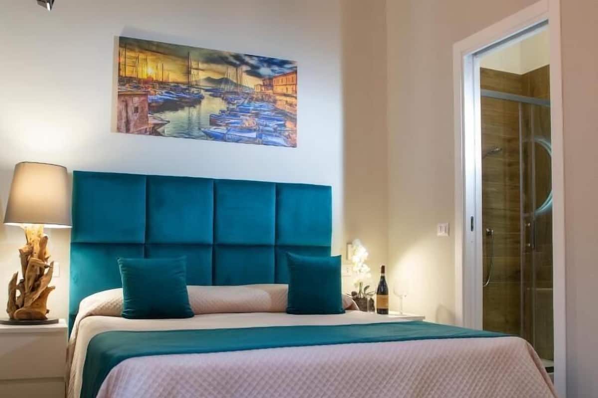 Um quarto com uma cama grande, cabeceira, mesa de cabeceira, abajur, um quadro na parede e porta do chuveiro de vidro.