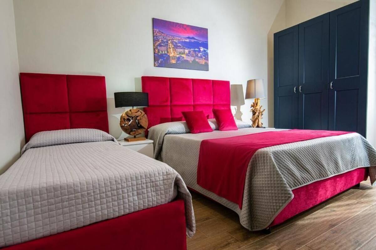 Quarto de hotel com duas camas, cada uma com cabeceira vermelha, mesa de cabeceira com luminária e um quadro na parede.