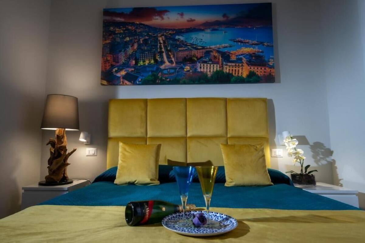 Quarto de hotel com cama, pintura de paisagem urbana, abajur e garrafa de champanhe.