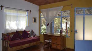 Une pièce avec un lit en bois, un bureau et une chaise. Il y a des fenêtres avec des rideaux, une porte et un ventilateur de plafond.