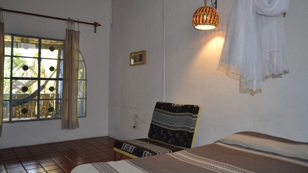 Une pièce comprenant un lit, une chaise, une serviette suspendue, une lampe et une fenêtre avec des rideaux.