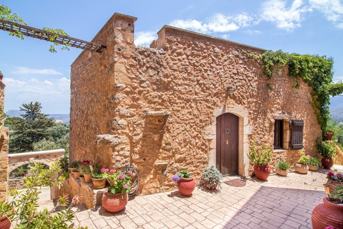 Edifici de pedra rústic amb porta i finestres de fusta, envoltat de plantes en test i un cel serè.