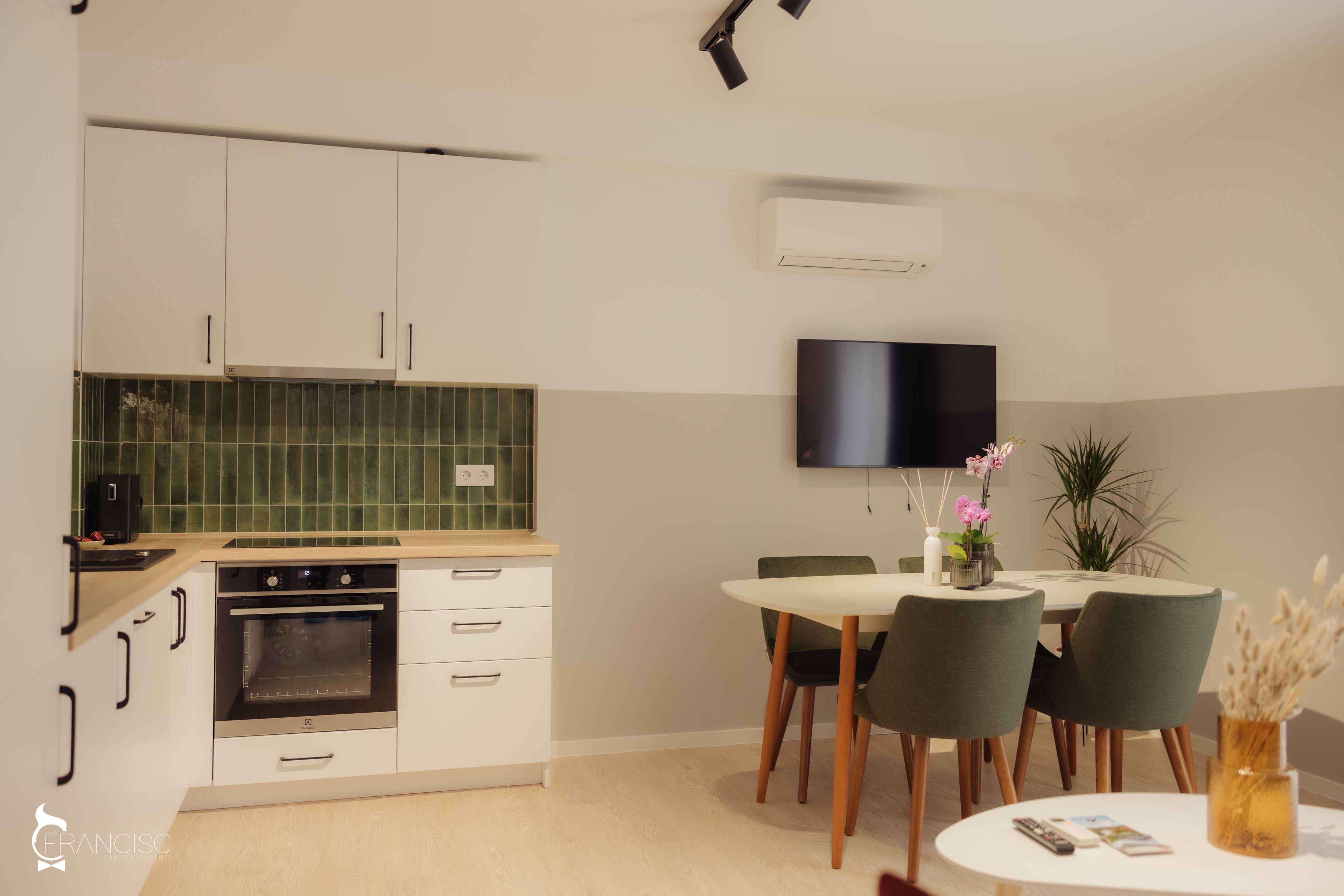A modern kitchen with white cabinets, a built-in oven, and a green backsplash. There is a dining area with a table and chairs, and a flat-screen TV mounted on the wall.