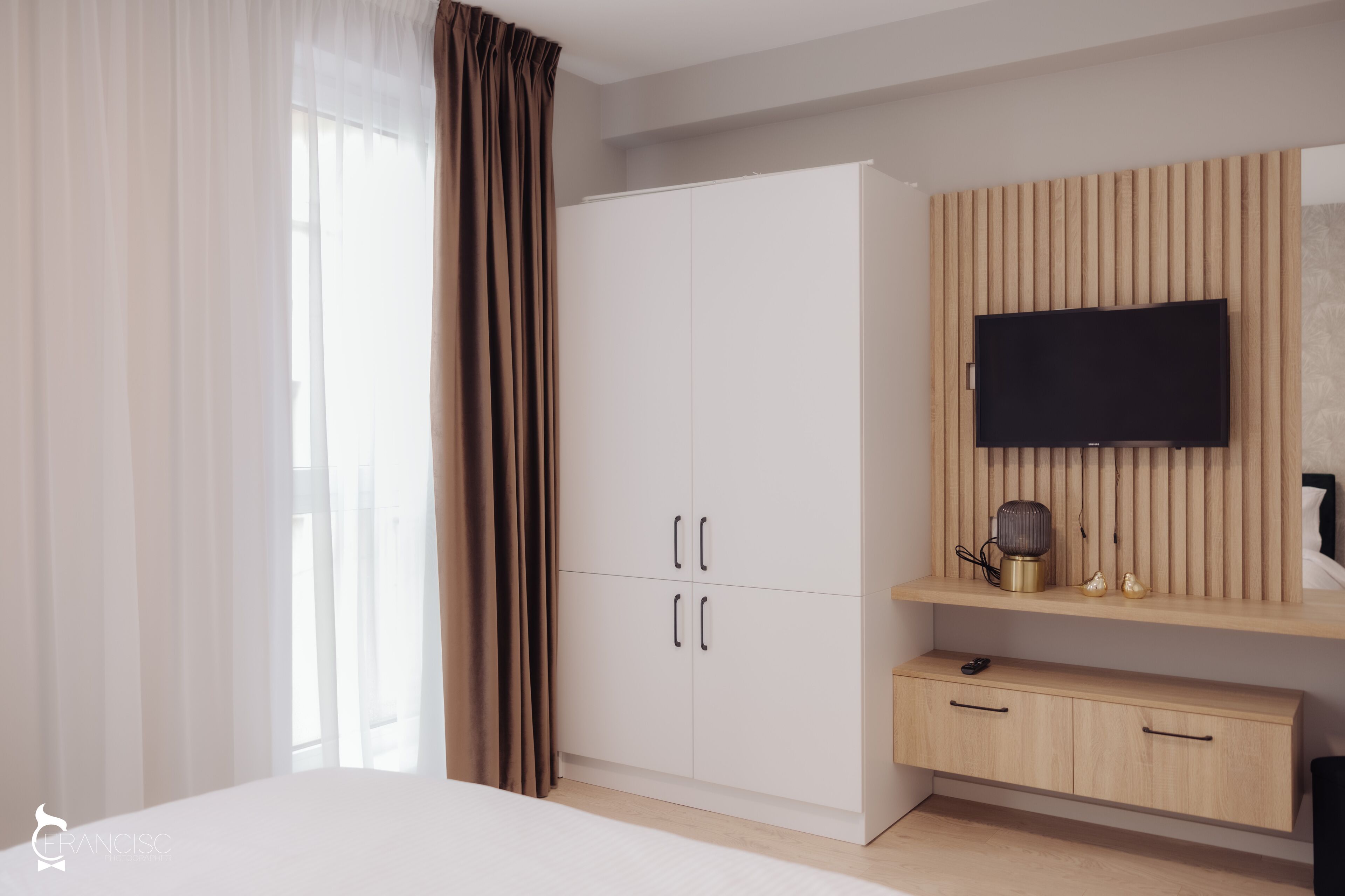 A bedroom with a bed, a wardrobe, a TV mounted on a wooden panel, and a bedside table with a lamp.
